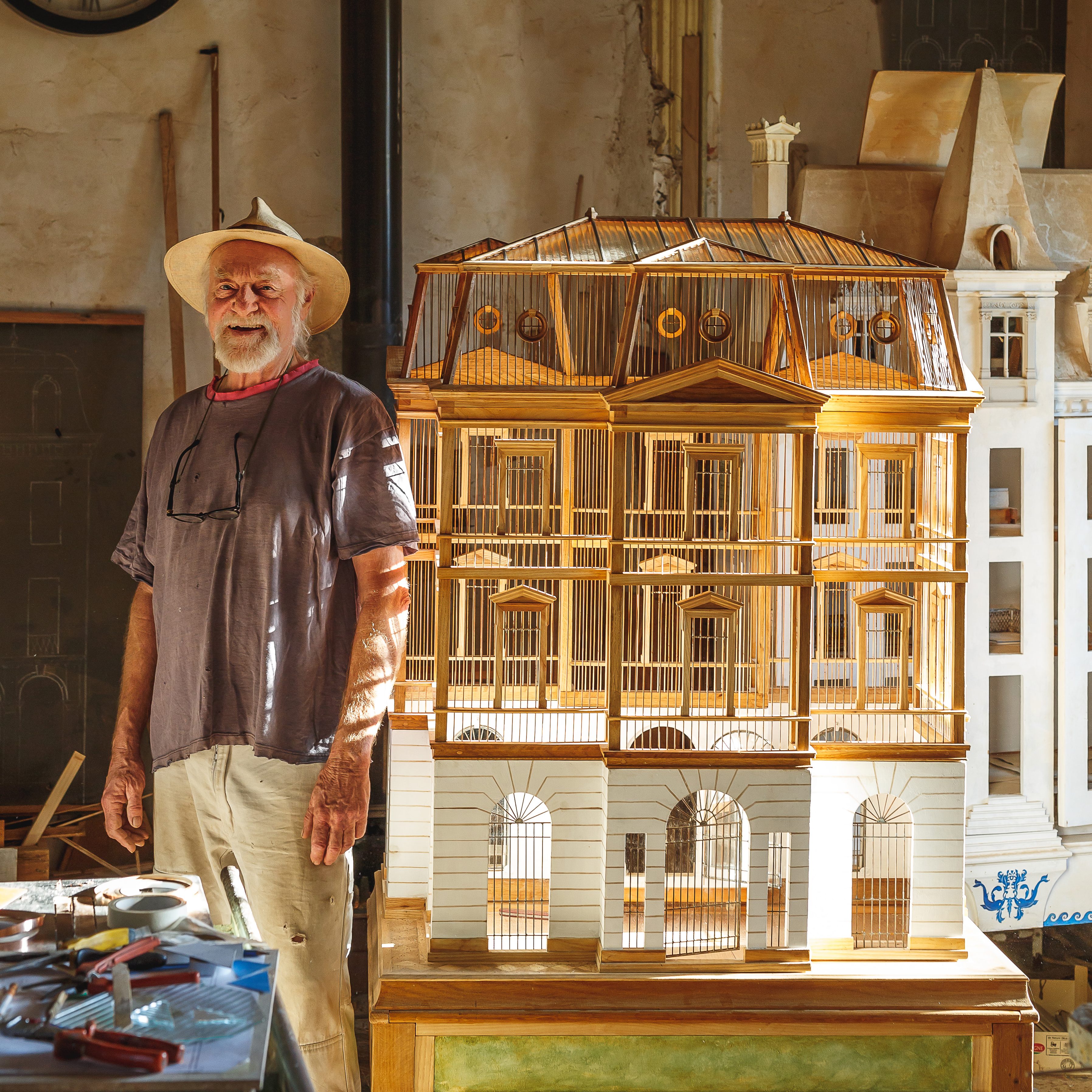 Eric wearing a hat and glasses resting on chest beaming in front of wooden birdcage. Birdcage rests on wood table with green accents and in front of natural stone wall